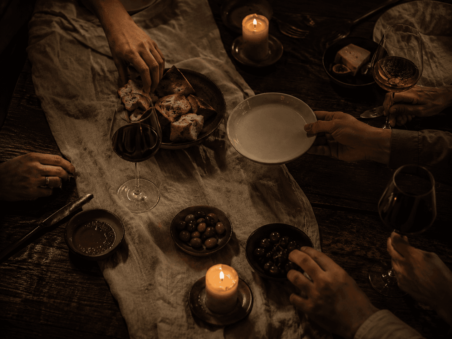 Hands reaching across a candlelit table for bread, olives and wine.